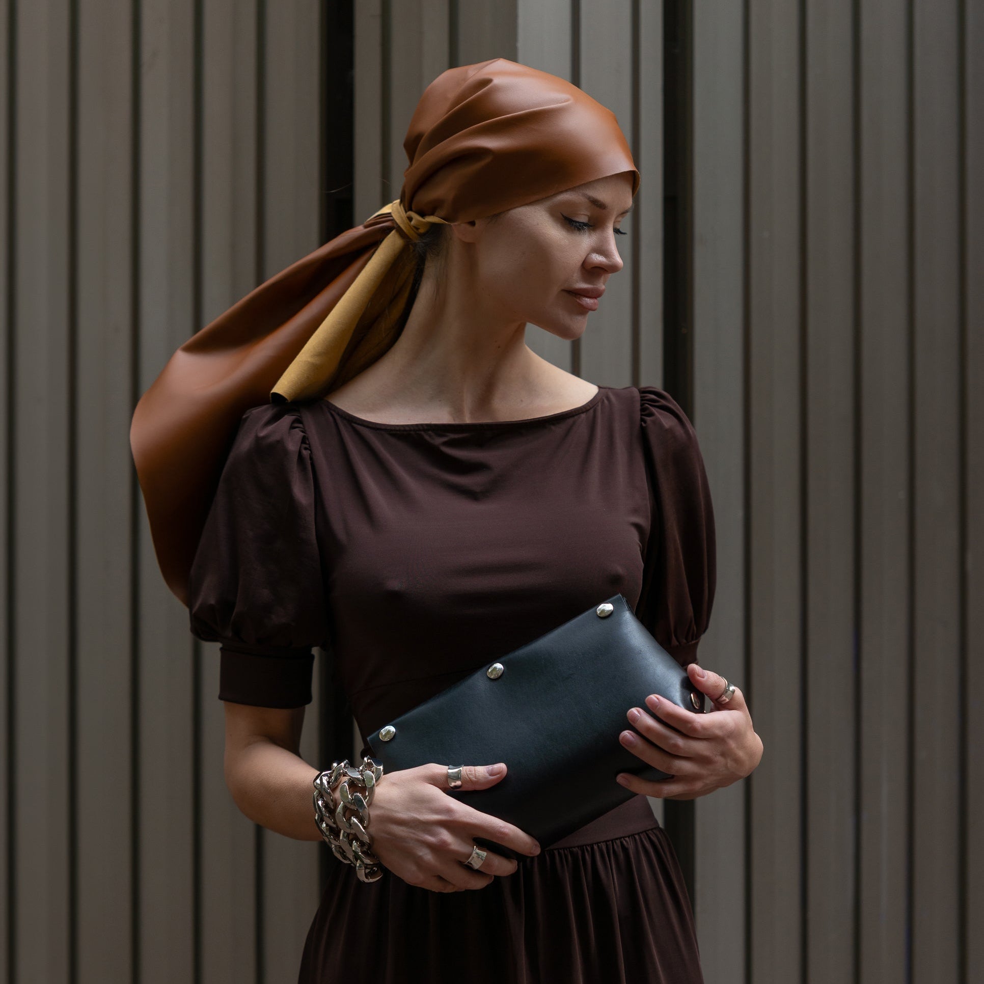 Woman in a brown dress with a brown headscarf holding a blue clutch against a gray background