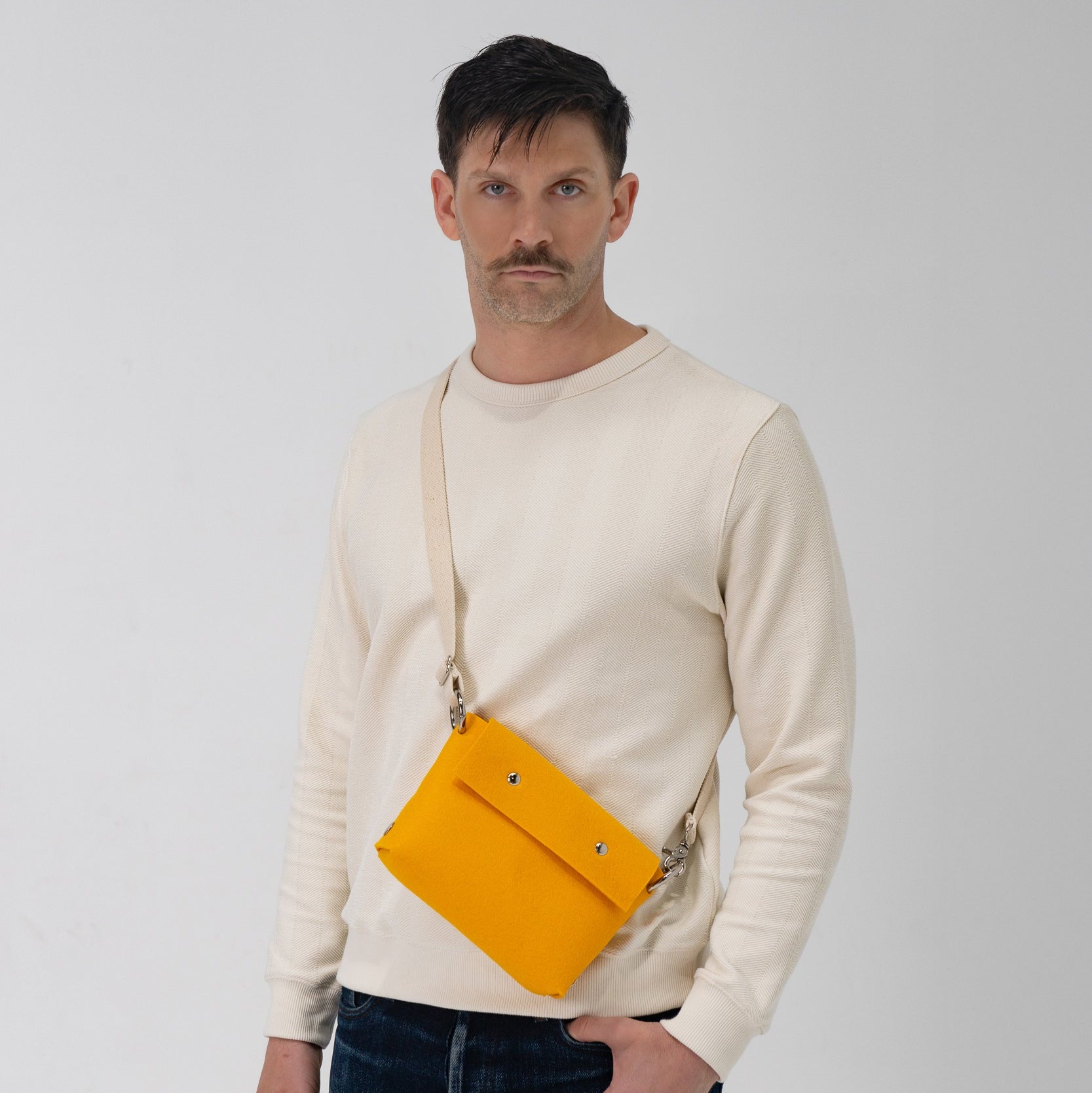 Man wearing a beige sweater and blue jeans with a yellow crossbody bag on a white background