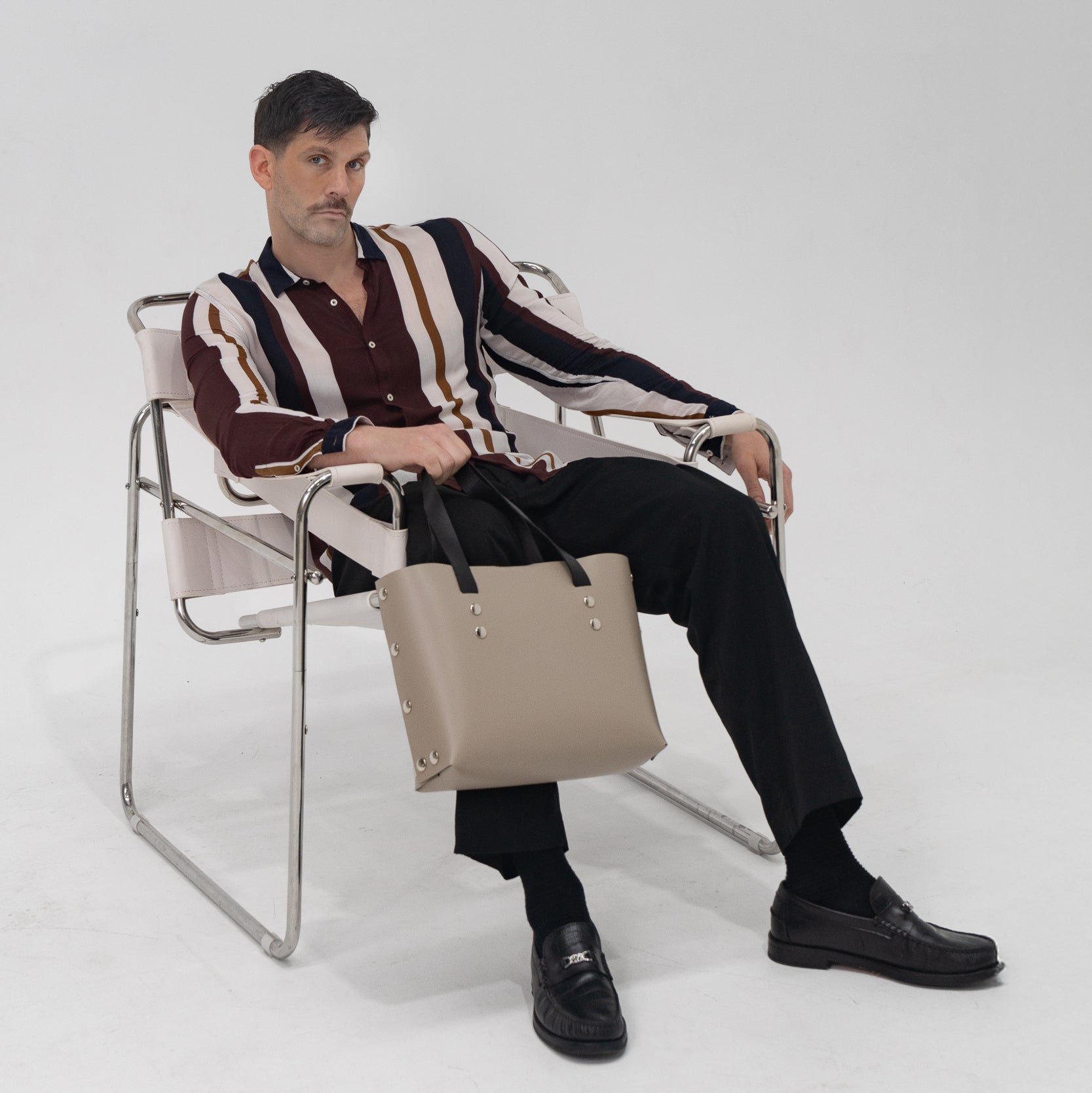 Man sitting on a chair holding a beige handbag against a white background