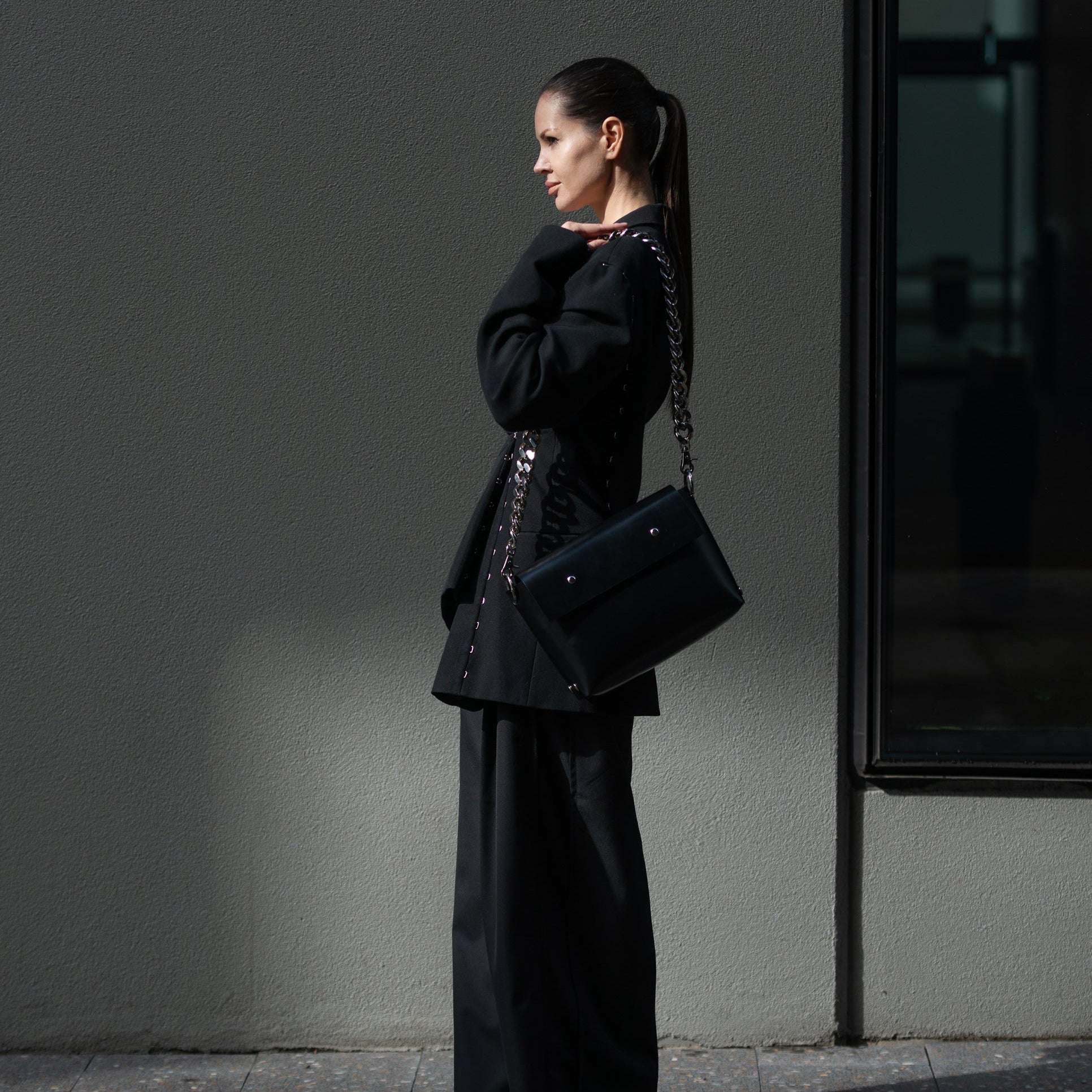 Woman in black outfit with a handbag standing against a gray wall.