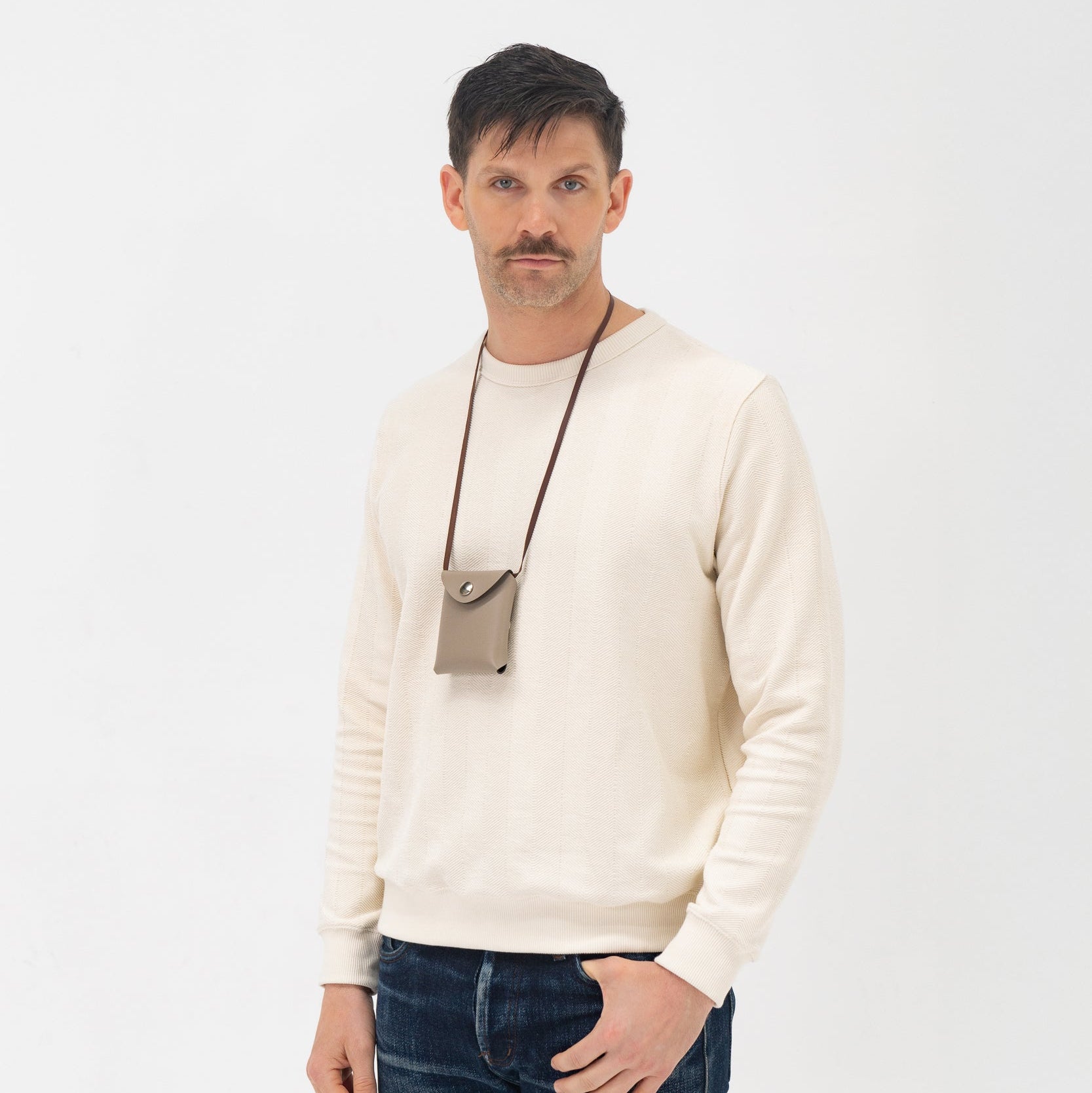 Man wearing a cream sweater and dark jeans with a neutral-colored bag over his shoulder on a white background