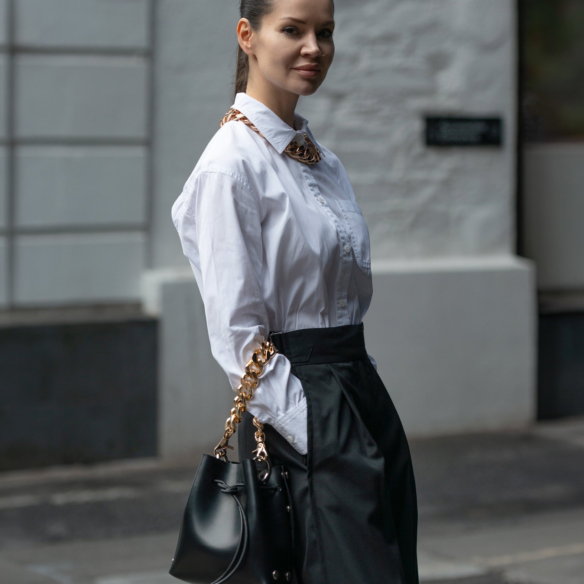 A woman in a white button-up shirt and black skirt holding a black handbag with a gold chain strap, standing outdoors against a light gray building wall.