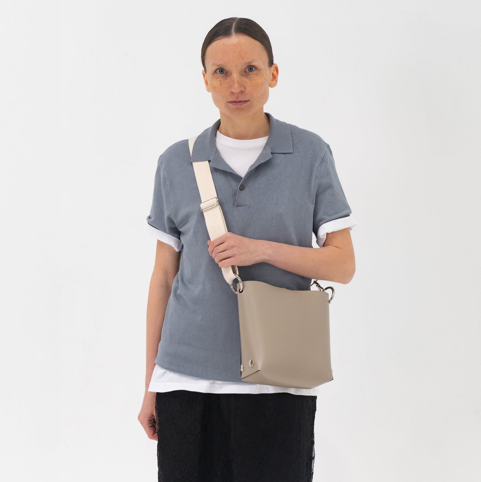 Person wearing a gray polo shirt and black skirt holding a beige bag on a white background